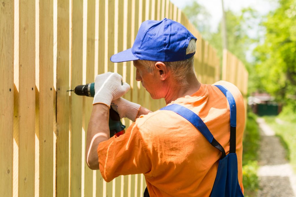 A picture of a man installing fence boards with a power drill.